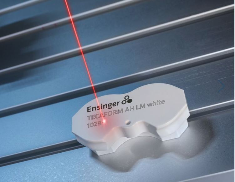 A close-up of a laser marking on a white plastic rod. The marking is black and reads "Ensinger\nTECAFORM AH LM white\n1028".