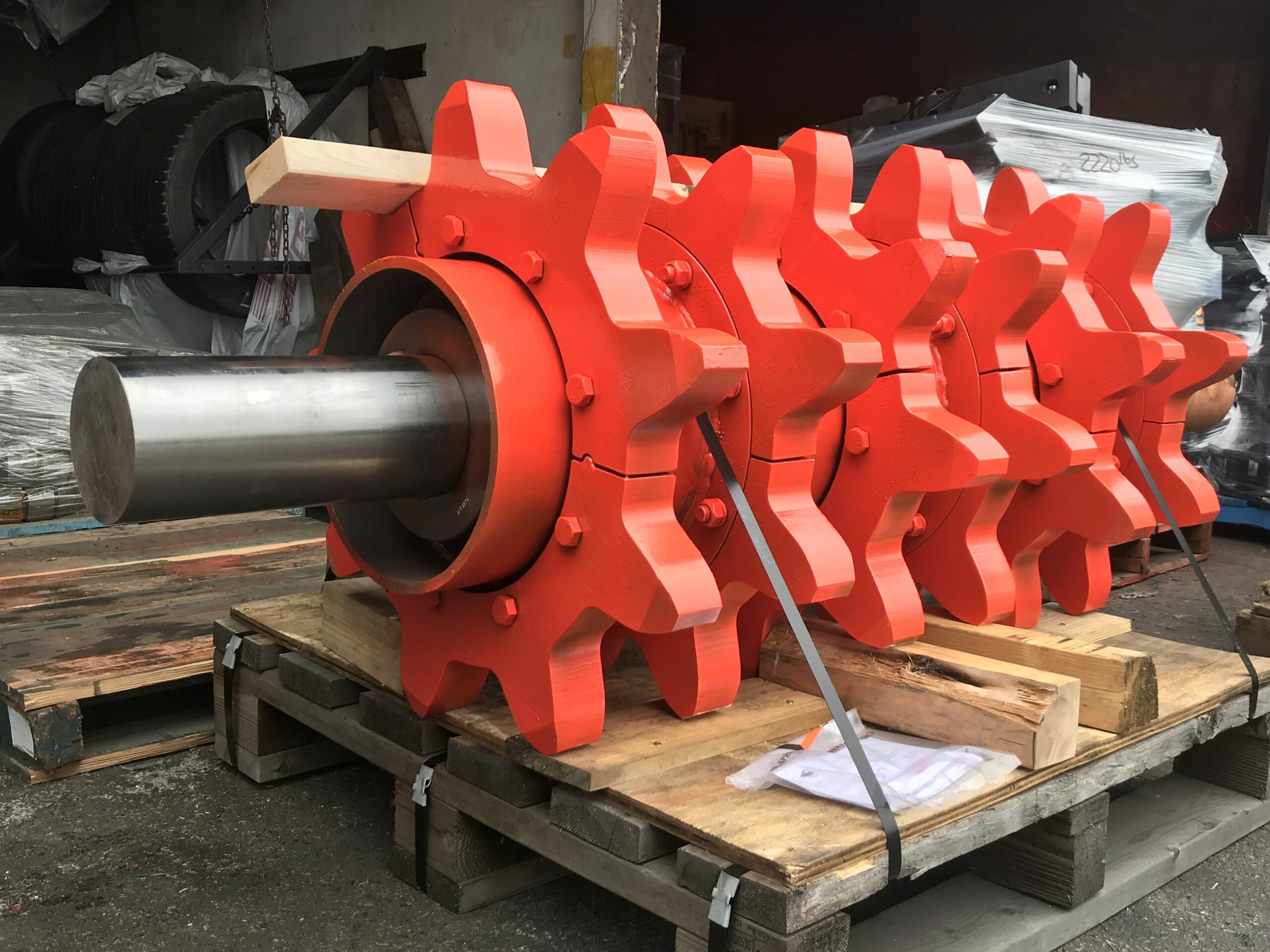 Close-up of a large, orange industrial machine with multiple visible gears, sitting on a wooden pallet in a factory setting.