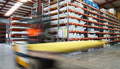 A yellow forklift carefully navigates through a busy warehouse with tall metal shelves stacked high with various supplies.