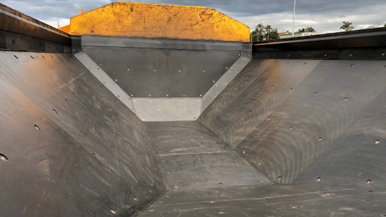 A close-up of the back of a dump truck with a raised hydraulic bed. The bed is empty and there is a metal chute in the middle.