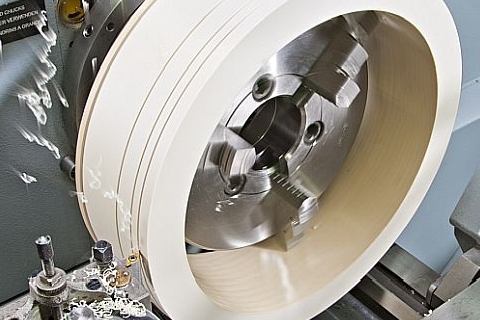 A close-up of a metal lathe with a rotating white cylinder being cut by a sharp tool.