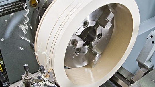 A close-up of a metal lathe with a rotating white cylinder being cut by a sharp tool.